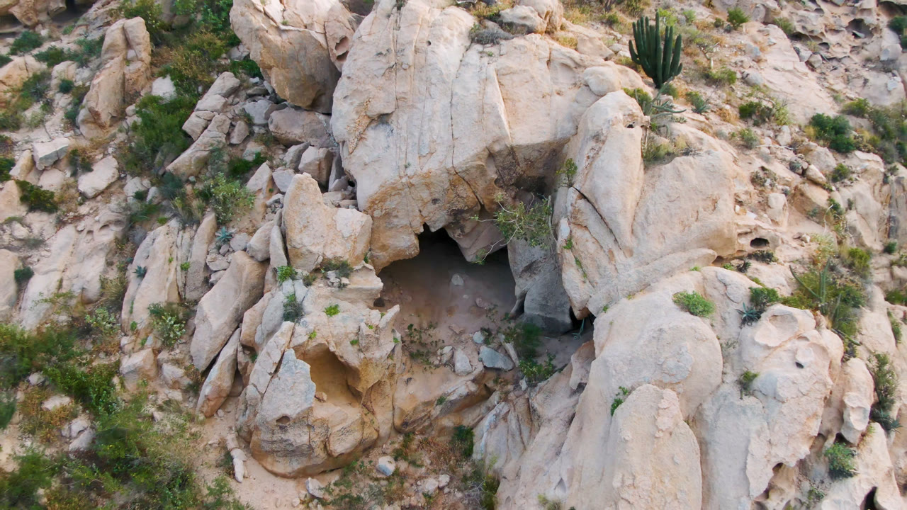 Aerial frame over Cabo Pulmo, Baja California Sur showing pale boulders, a dark cave opening near the lower left, sparse green shrubs, cactus, and no visible people.
