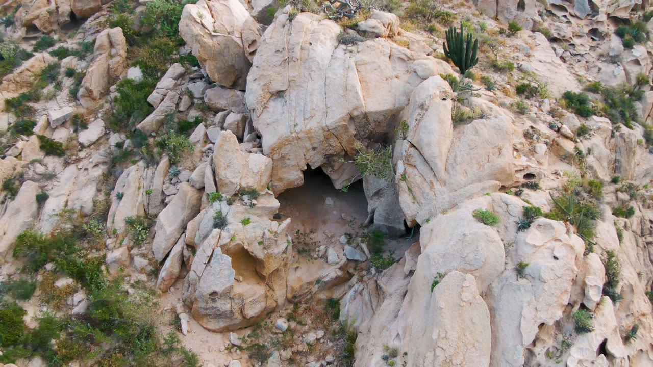Overhead frame in Cabo Pulmo, Baja California Sur with the cave mouth lower left of center, tall pale rock slabs, scrub vegetation, cactus, and no visible people.