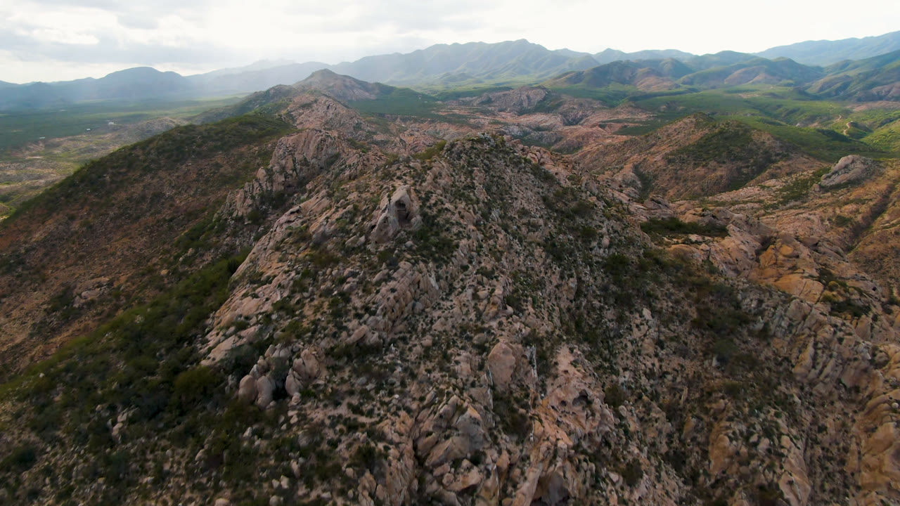 Aerial frame over Cabo Pulmo, Baja California Sur, showing a rocky ridge in the foreground, green scrub on the slopes, distant mountains, and no visible people.
