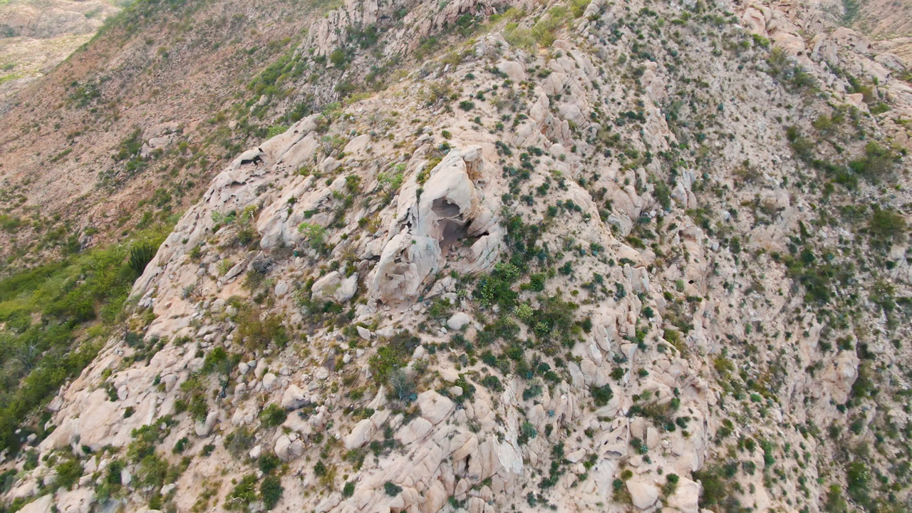 Opening frame of a rocky Cabo Pulmo hillside seen from above, with a dark cave opening in pale stone and no visible people.