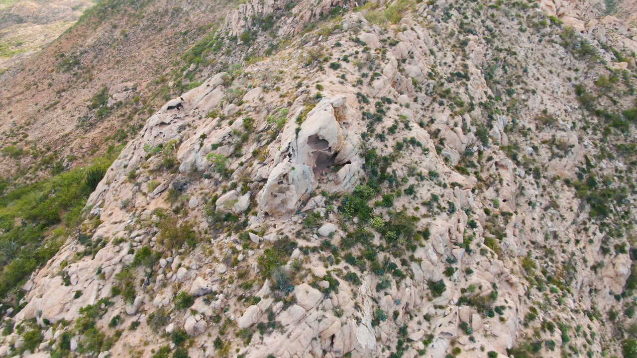 Aerial frame over Cabo Pulmo terrain with the cave feature slightly left of center, pale fractured rock, scattered green vegetation, and no visible people.
