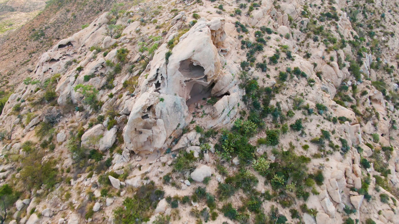 Aerial frame over Cabo Pulmo, Baja California Sur, showing a small dark cave opening in pale rounded rock, scattered green shrubs, sandy terrain, and no visible people.