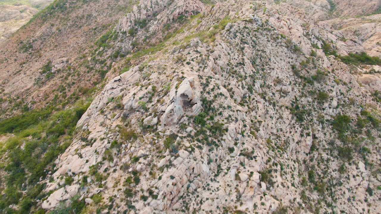 Aerial frame from Cabo Pulmo, Baja California Sur, with the boulder-strewn ridge centered, green brush in the gullies, a small perched bird, and no visible people.