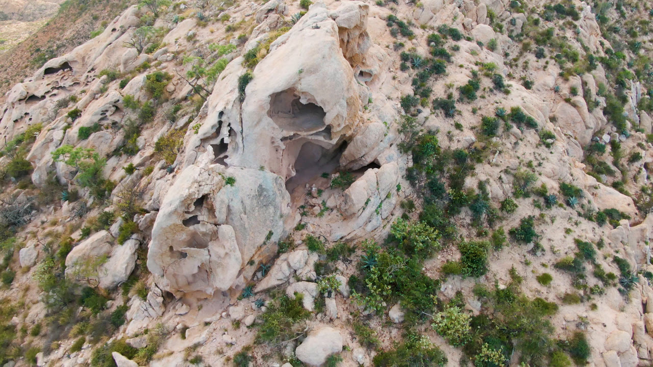 Aerial frame from Cabo Pulmo, Baja California Sur, showing a pale tan rock formation with a dark cave opening, sandy slopes, green shrubs, and no visible people at the start of the clip.