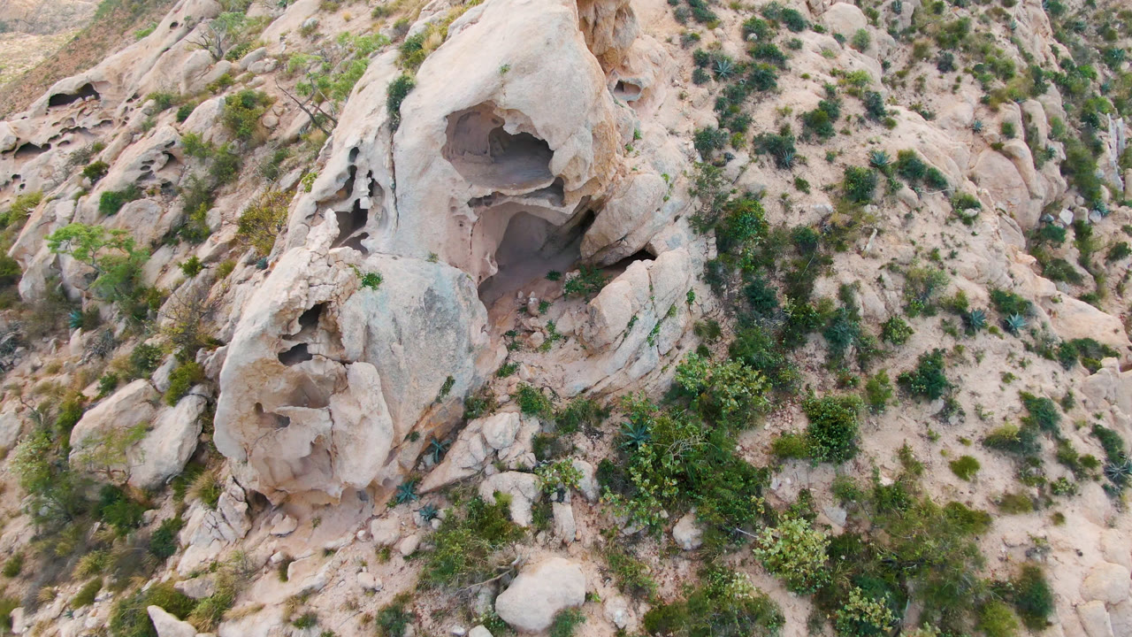 Drone perspective on a large hollowed rock surrounded by dry hillside plants