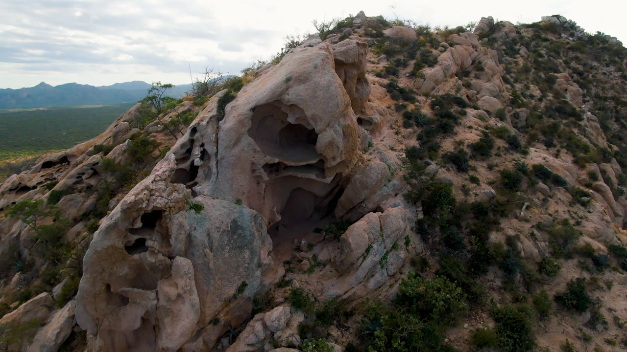 Large weathered rock with dark cave openings rises above scrubby hillside terrain in Cabo Pulmo, Baja California Sur, with distant mountains and no visible people.