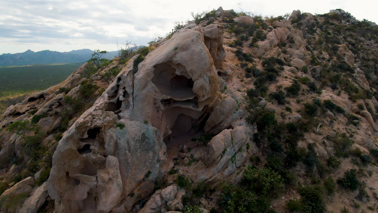 The cave-like rock face sits left of center in Cabo Pulmo, Baja California Sur, with layered tan stone, sparse shrubs, distant mountains, and no visible people.