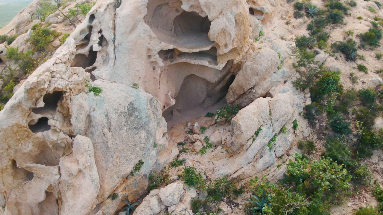 Opening frame over pale eroded rock in Cabo Pulmo, Baja California Sur, with dark cave pockets, sandy ground, scattered shrubs, and no visible people.
