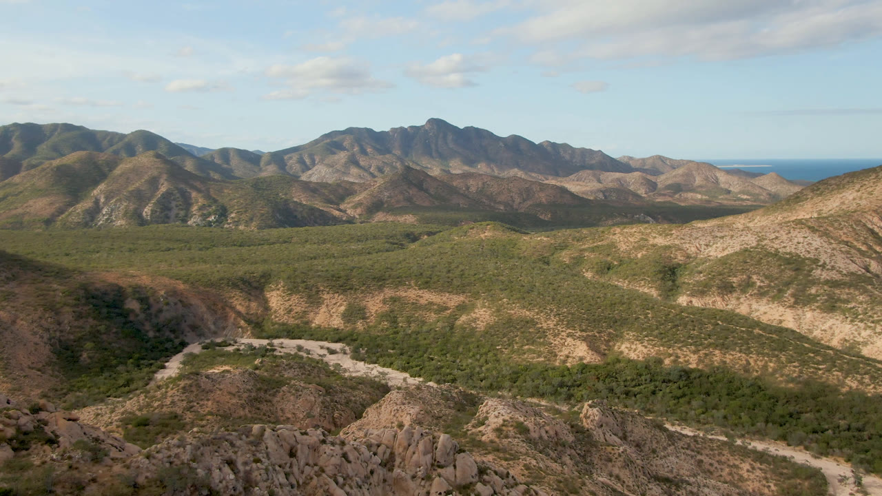 Opening frame over Cabo Pulmo, Baja California Sur, with rocky foreground ridges, a green valley, a pale road, distant mountains, blue ocean on the horizon, and no visible people.