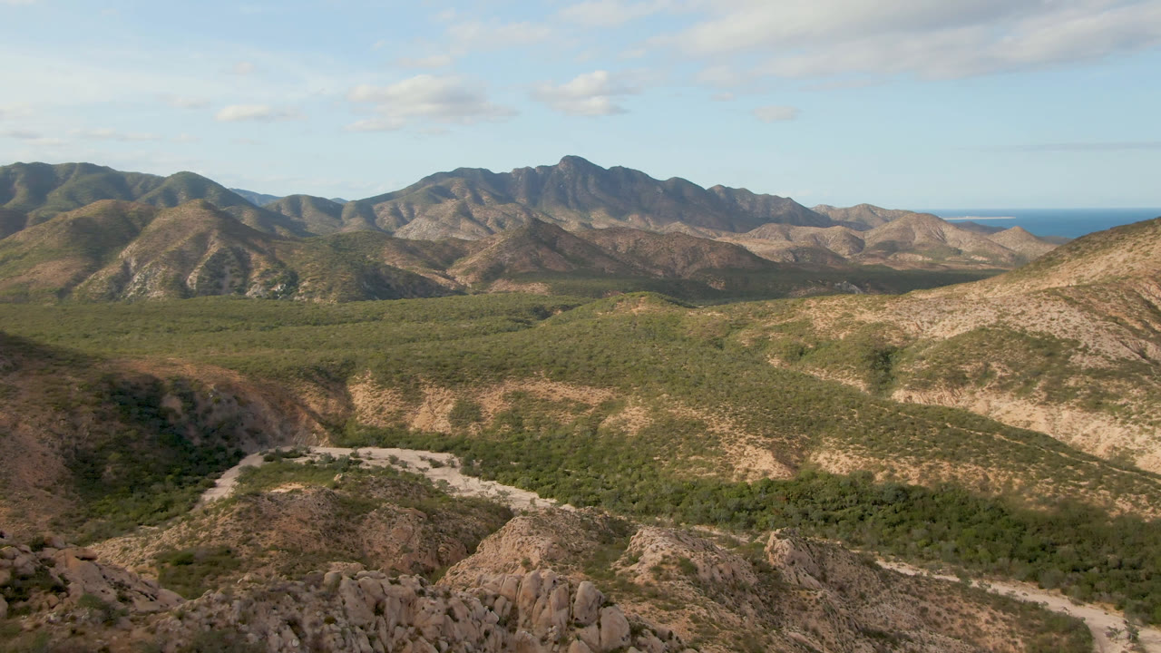 Second frame from the Cabo Pulmo aerial view, with the winding road more centered through green scrub, layered mountain slopes behind it, distant ocean, and no visible people.