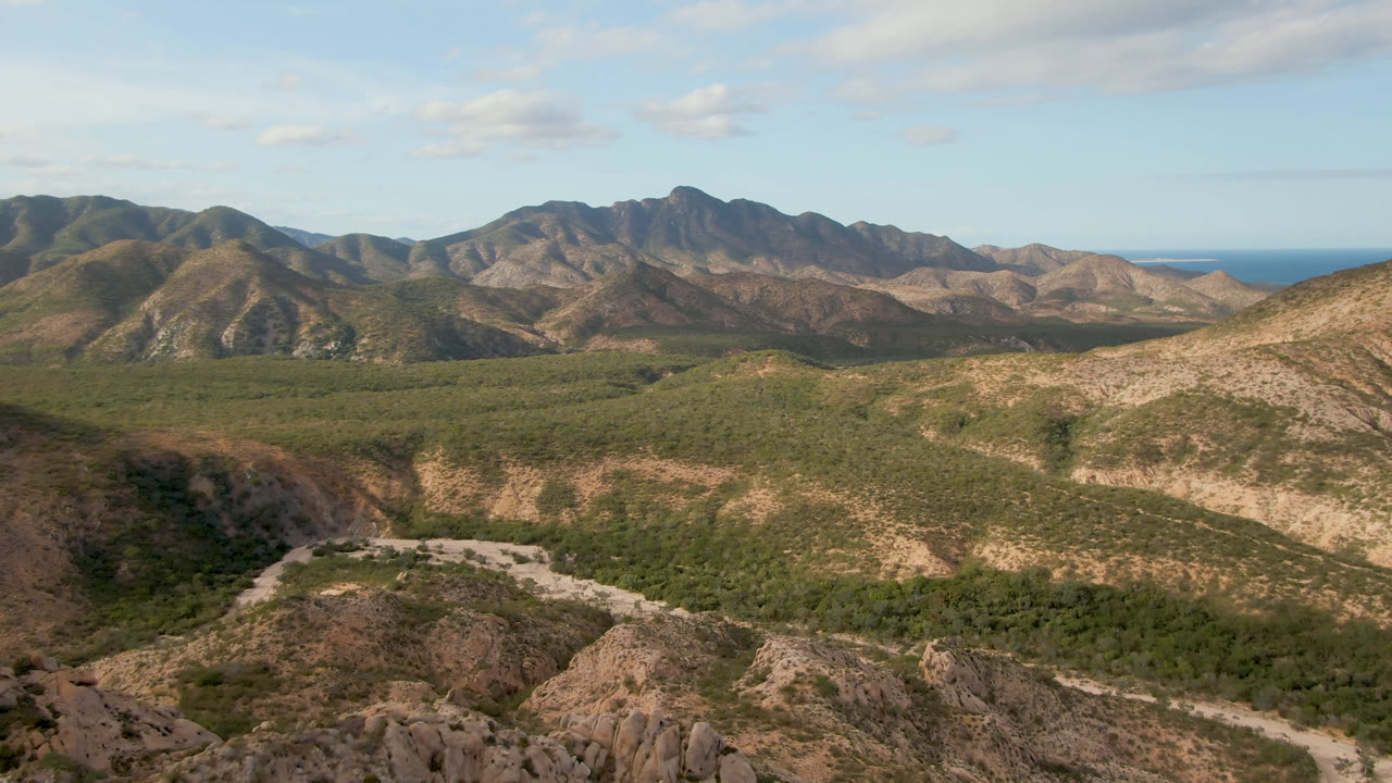 Rocky desert foreground above a green valley, winding road, mountains, and ocean horizon.
