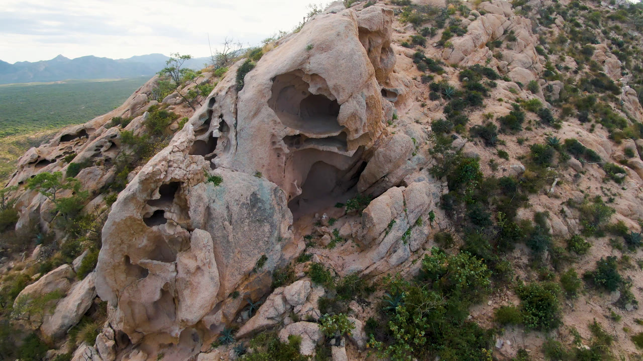 A close aerial view in Cabo Pulmo, Baja California Sur, shows a pale eroded rock formation with several hollow openings, desert shrubs below, distant mountains on the horizon, and no visible people.