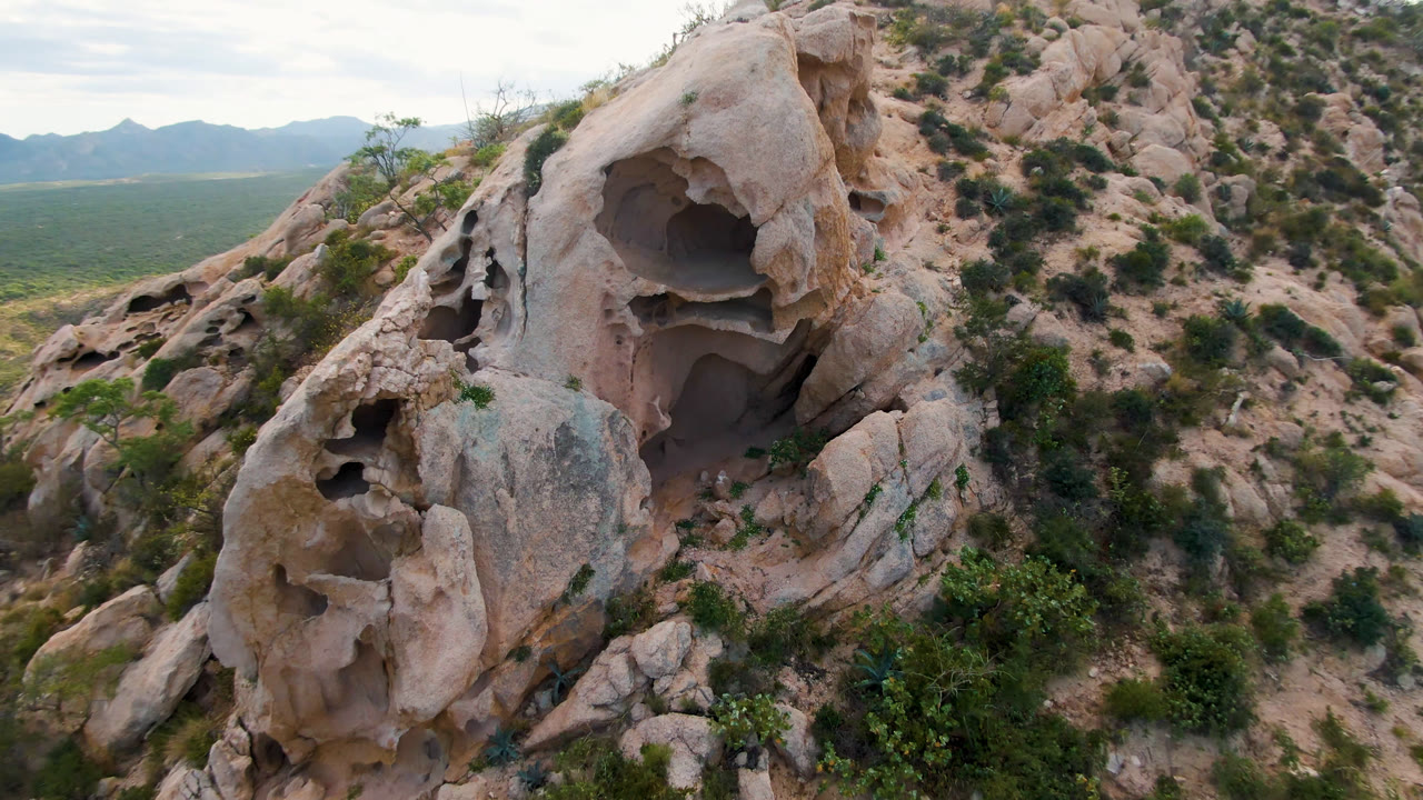 Aerial framing in Cabo Pulmo centers the large upper hollow in the pale rock, with smaller cavities along the left side, green shrubs at the base, mountain silhouettes behind, and no visible people.