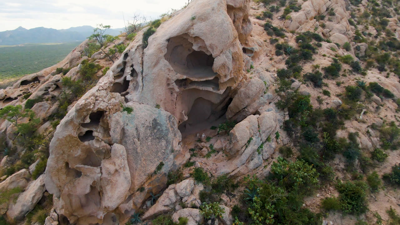 Eroded rock cave face beside desert brush with a low mountain horizon.