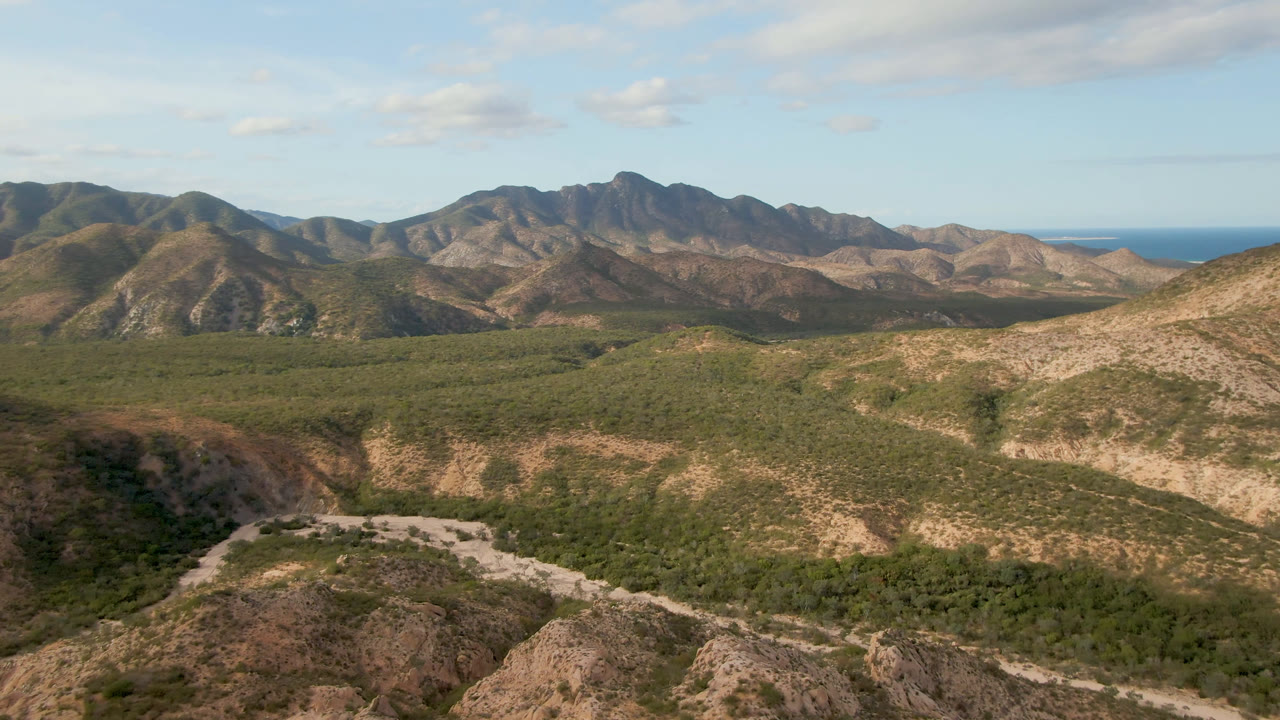 Opening frame over Cabo Pulmo shows tan desert hills, green scrub, a pale road curving through the valley, distant mountains, ocean on the horizon, and no visible people.