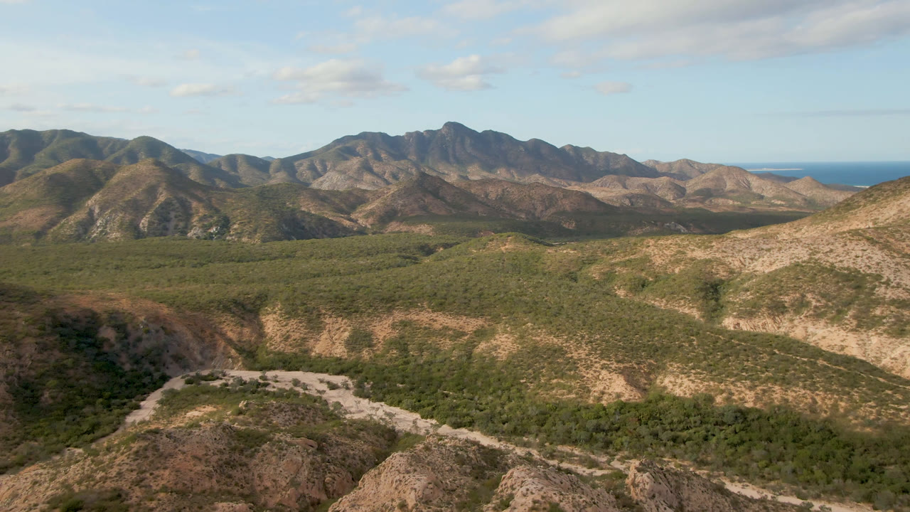 At one second, the Cabo Pulmo landscape centers the curving road and scrub-filled valley below layered brown mountains, distant ocean, light clouds, and no visible people.