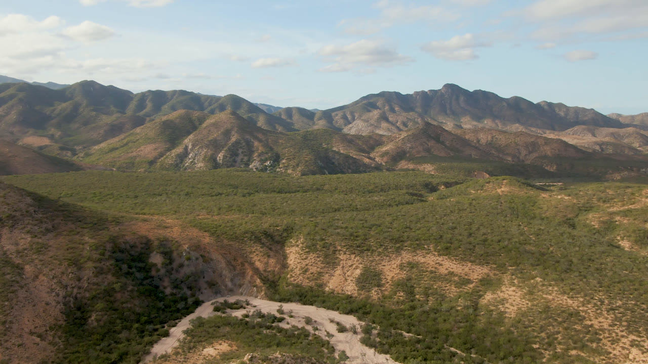 Wide aerial frame over Cabo Pulmo, Baja California Sur, showing green scrubland, tan rocky ridges, distant mountains, scattered clouds, and no visible people.