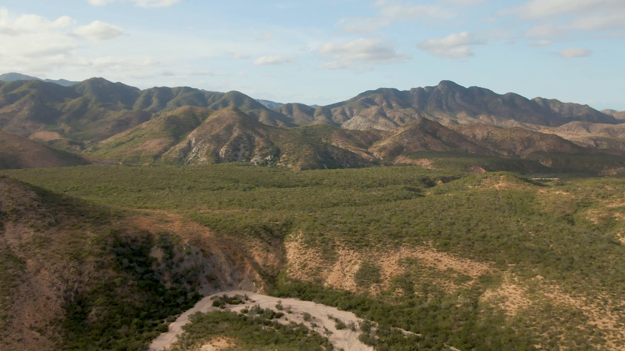 Aerial view in Cabo Pulmo with a foreground ridge, broad scrub-covered valley, pale road traces, layered mountain slopes, blue sky, and no visible people.