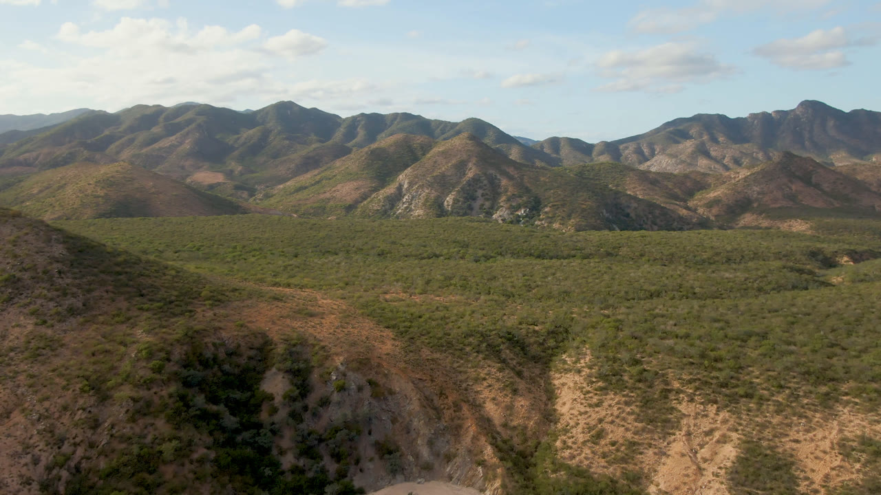 Opening aerial frame over Cabo Pulmo, Baja California Sur, showing tan foreground slopes, green valley vegetation, distant rugged mountains, blue sky, and no visible people.