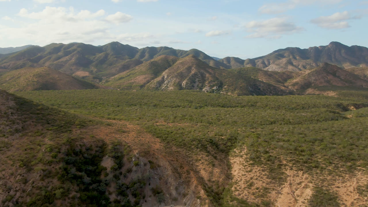 Second aerial frame in Cabo Pulmo with the green midground valley stretching below layered dry mountain ridges and a bright sky with scattered clouds, with no visible people.