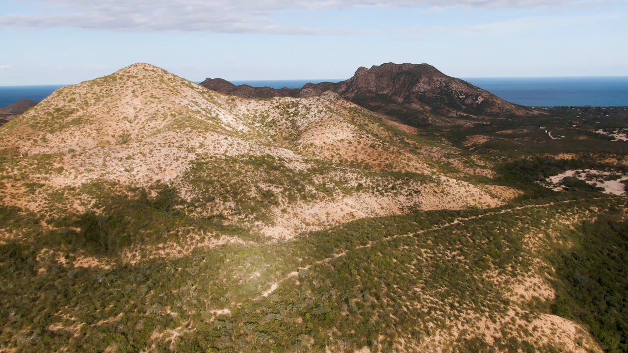 Cabo Pulmo aerial frame showing a rounded tan mountain in the foreground, darker rock formations to the right, distant blue ocean, and no visible people.