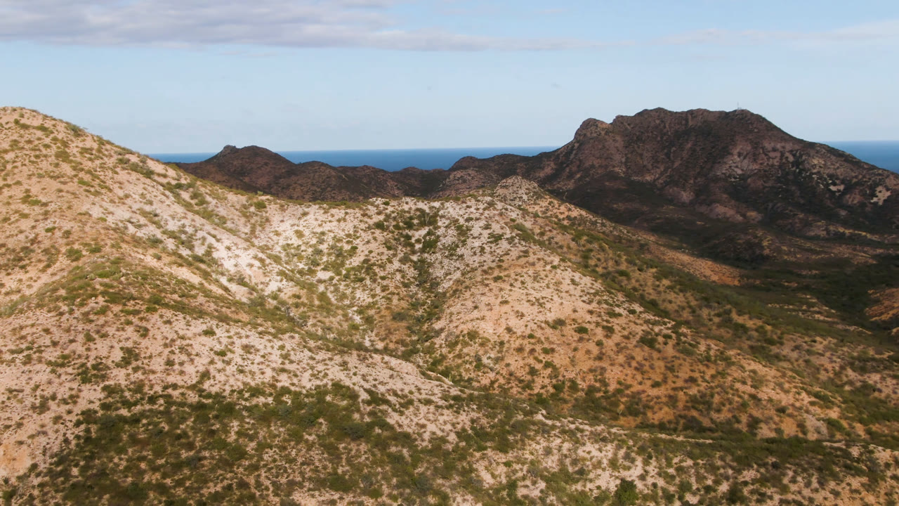 Opening aerial frame over Cabo Pulmo, Baja California Sur, with pale scrub hills in front, dark rocky mountains behind, blue ocean on the horizon, and no visible people.