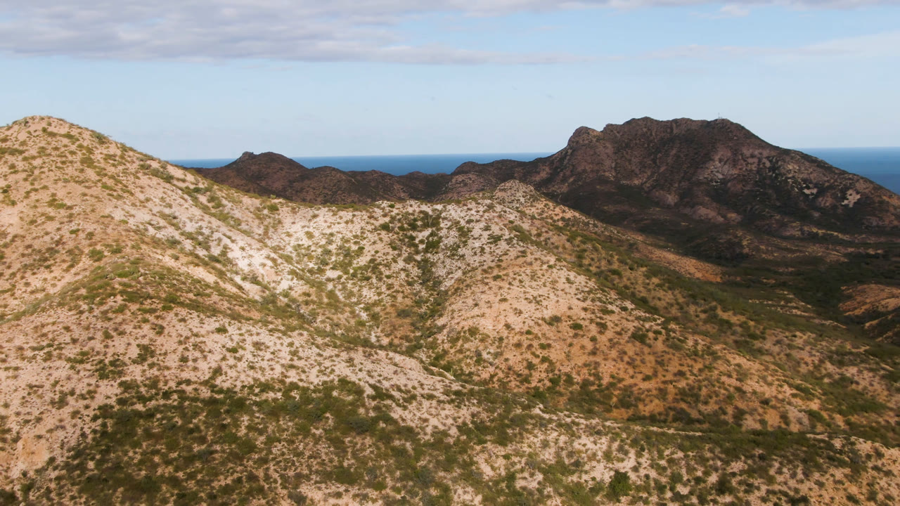 Aerial frame in Cabo Pulmo showing sunlit tan hills with sparse green vegetation, a shadowed mountain ridge, a thin blue ocean band, and no visible people.