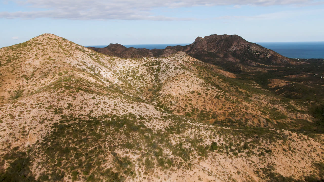 Aerial frame from Cabo Pulmo, Baja California Sur, showing a pale scrub-covered hill in the foreground, darker mountain ridges behind it, a blue ocean horizon, and no visible people.