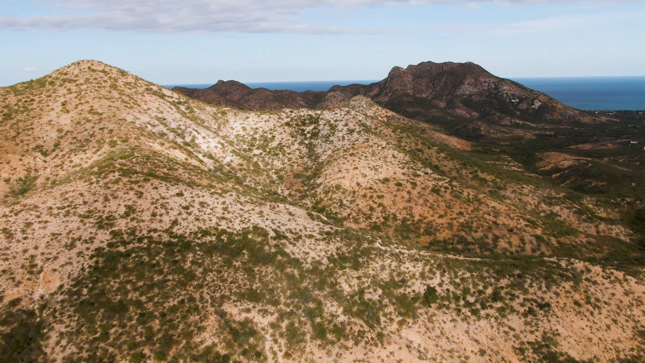 Aerial view in Cabo Pulmo with the foreground hill shifted slightly across the frame, showing scrubby tan slopes, dark ridges, distant ocean, blue sky, and no visible people.