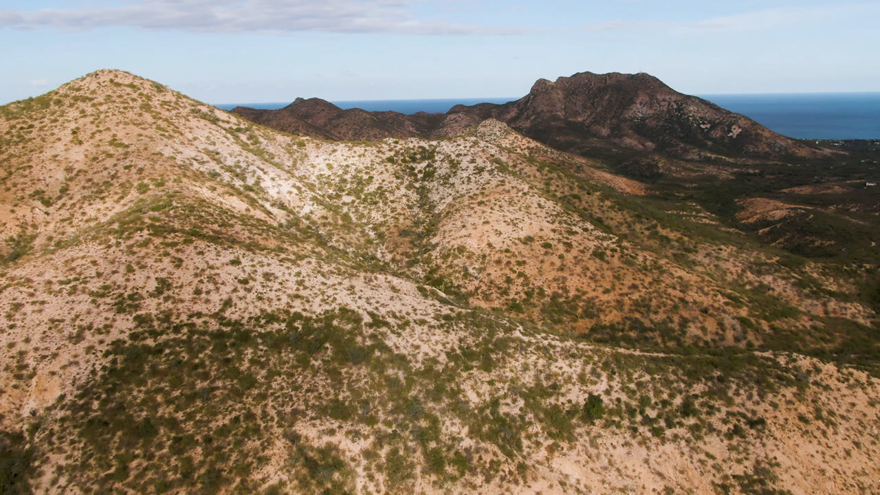 Desert mountain slope with sparse vegetation, distant peaks, and ocean horizon.