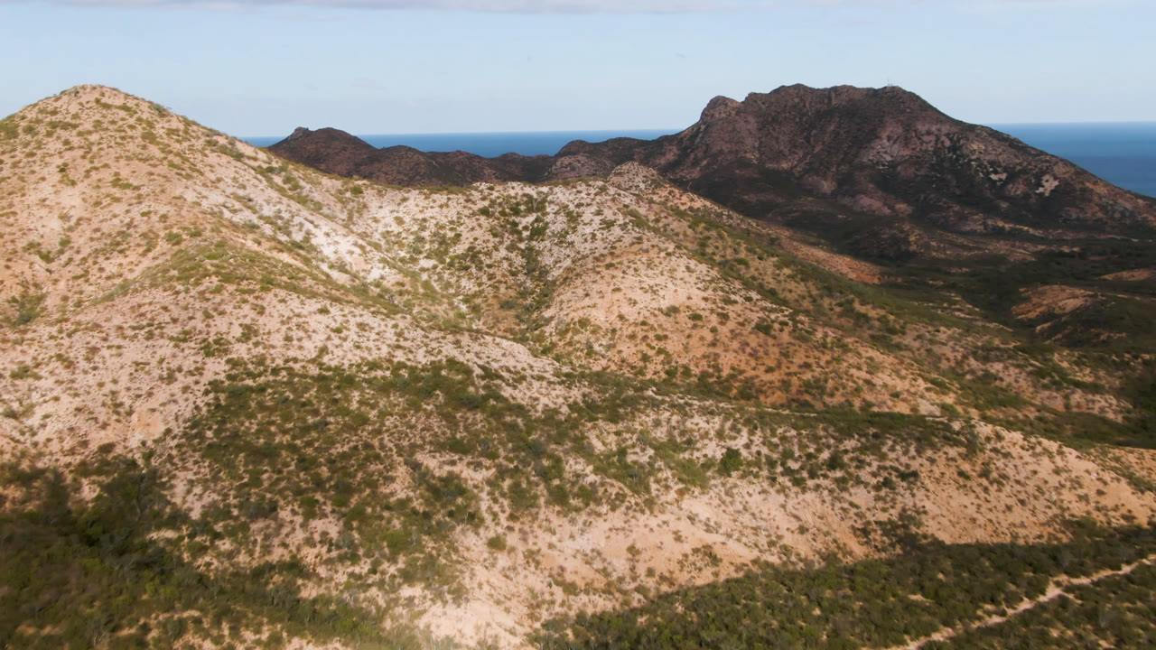 Aerial frame over Cabo Pulmo showing pale scrub-covered ridges in front of darker rocky mountains, with a blue ocean horizon and no visible people.