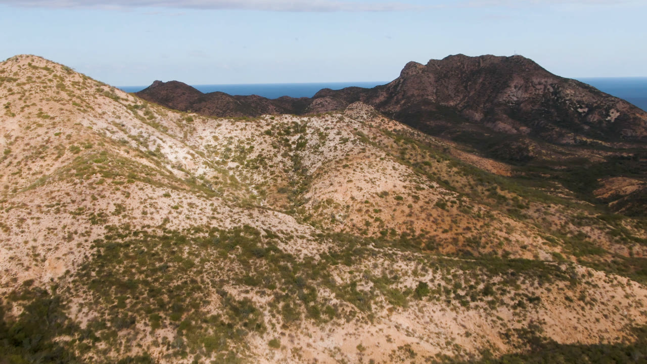 Cabo Pulmo aerial view with a sunlit ridge running across the foreground, darker mountain mass behind it, ocean horizon near the top, and no visible people.