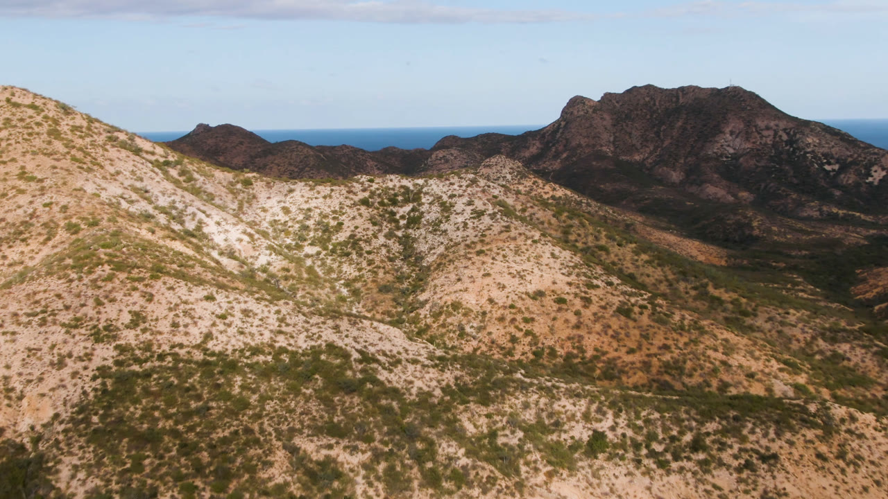 Wide aerial landscape of arid ridges, dark mountains, and a blue sea horizon.