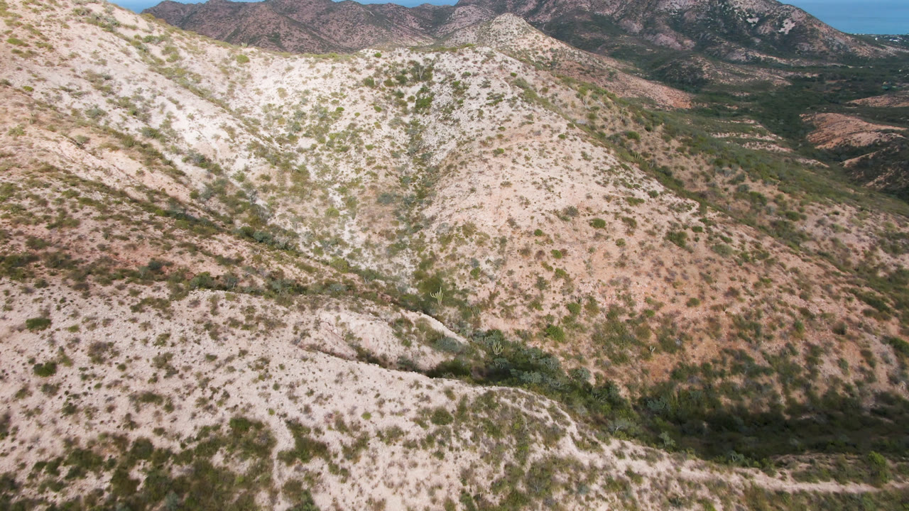Opening aerial frame over pale rocky hills and scrub-covered slopes in Cabo Pulmo, Baja California Sur, with distant mountain ridges and no visible people.