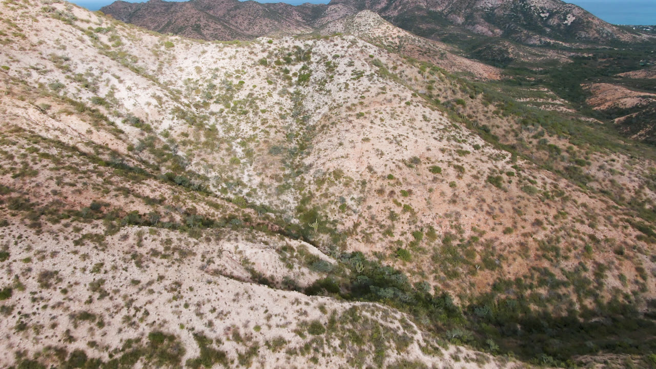 Aerial frame in Cabo Pulmo showing scrub-dotted rocky slopes, a shallow gully through the hills, darker ridges in the background, and no visible people.