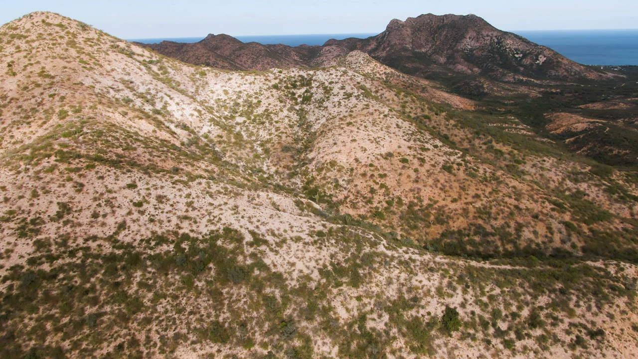 Aerial view over Cabo Pulmo, Baja California Sur, with pale scrub-covered hills in front, darker mountains beyond, blue sea on the horizon, and no visible people.
