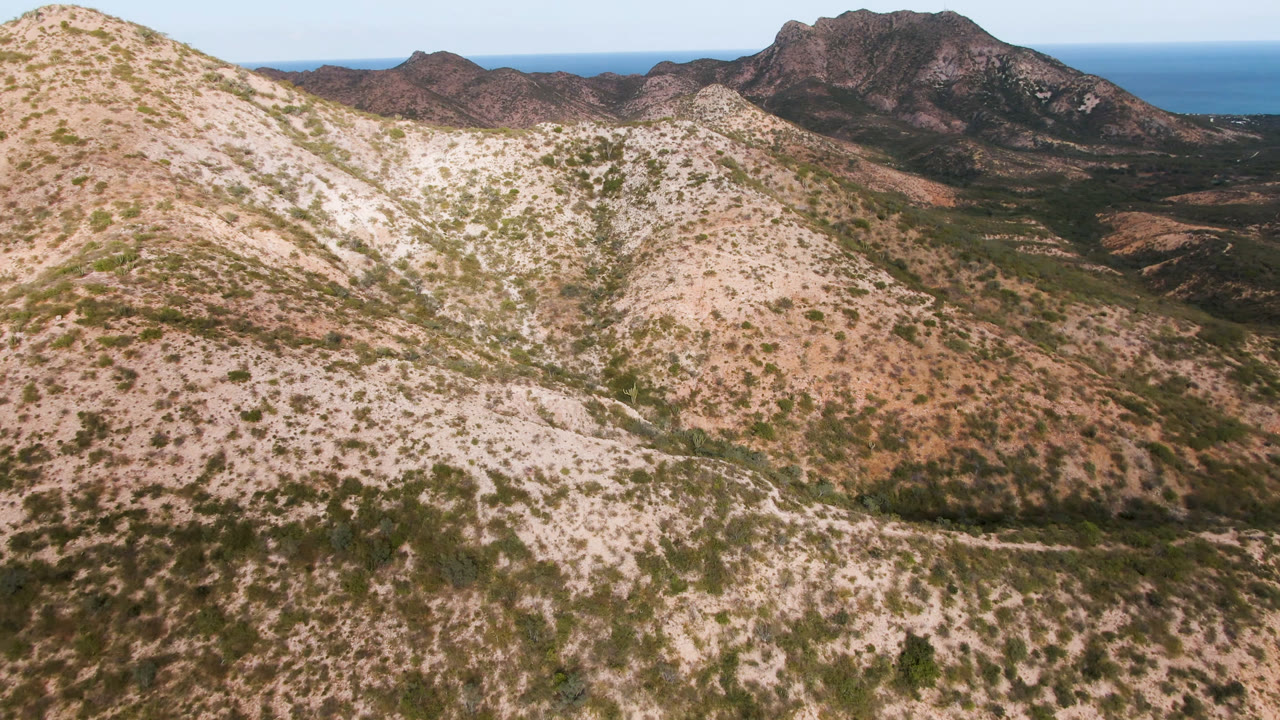 Elevated Cabo Pulmo landscape view with a pale foreground ridge, scattered shrubs, shadowed mountain ridges in the distance, a blue sea line, and no visible people.