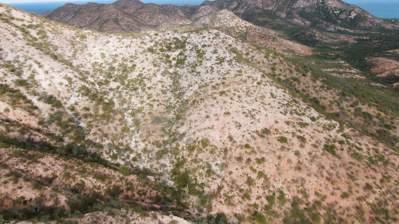 Opening aerial frame over Cabo Pulmo, Baja California Sur, showing a pale scrub-covered hillside, darker valley vegetation, rocky mountains, distant blue ocean, and no visible people.
