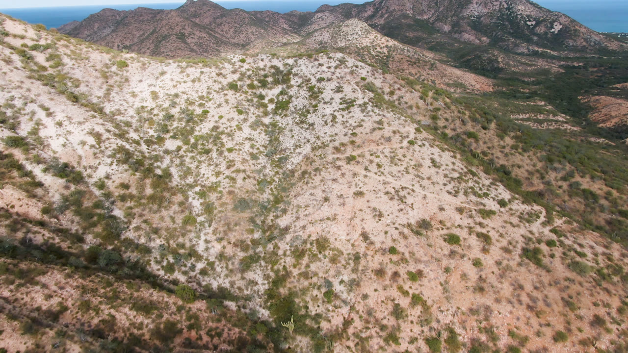 Aerial frame from Cabo Pulmo with the pale ridge filling the foreground, a dark green valley behind it, rugged mountains across the horizon, distant ocean, and no visible people.
