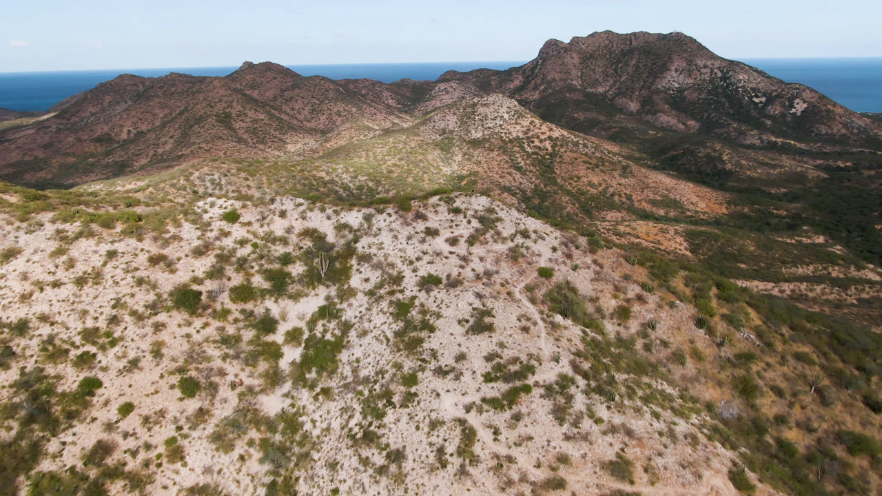 Aerial frame over Cabo Pulmo showing pale rocky foreground hills, scattered green shrubs, darker mountain ridges, blue ocean at the horizon, and no visible people.