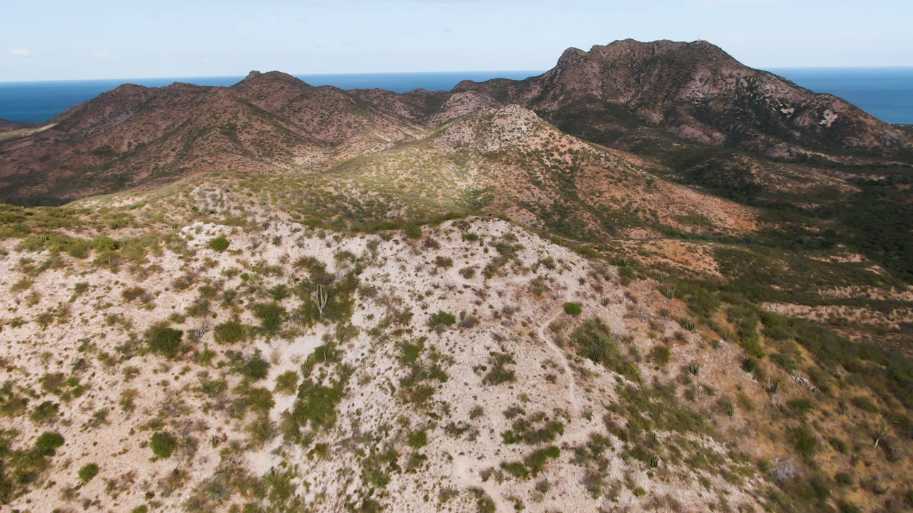 Aerial view in Cabo Pulmo with a pale shrub-dotted slope in front, darker ridges beyond, a prominent mountain mass to the right, blue ocean behind it, and no visible people.