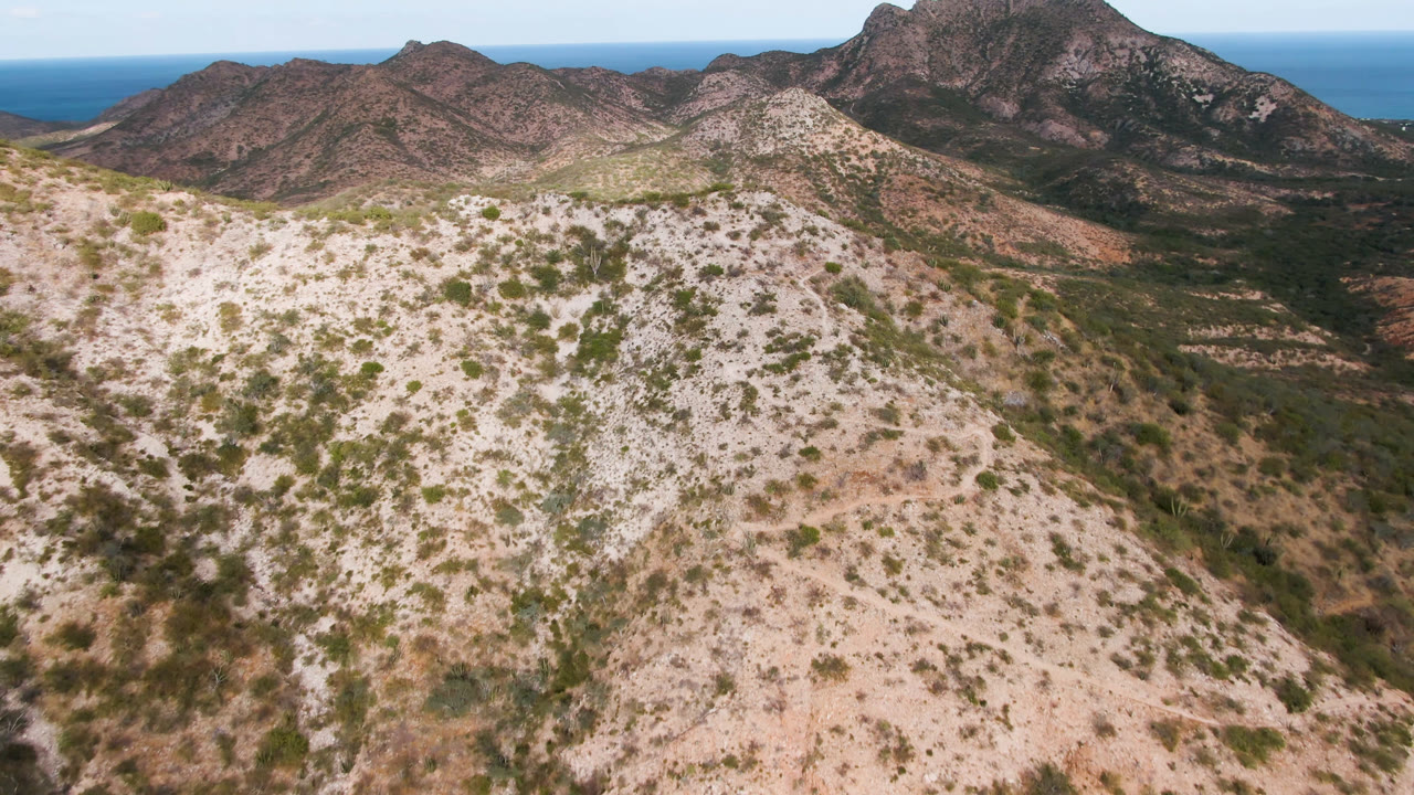 Aerial frame from Cabo Pulmo, Baja California Sur, Mexico showing pale rocky hills with sparse shrubs, darker mountains beyond, blue ocean on the horizon, and no visible people.