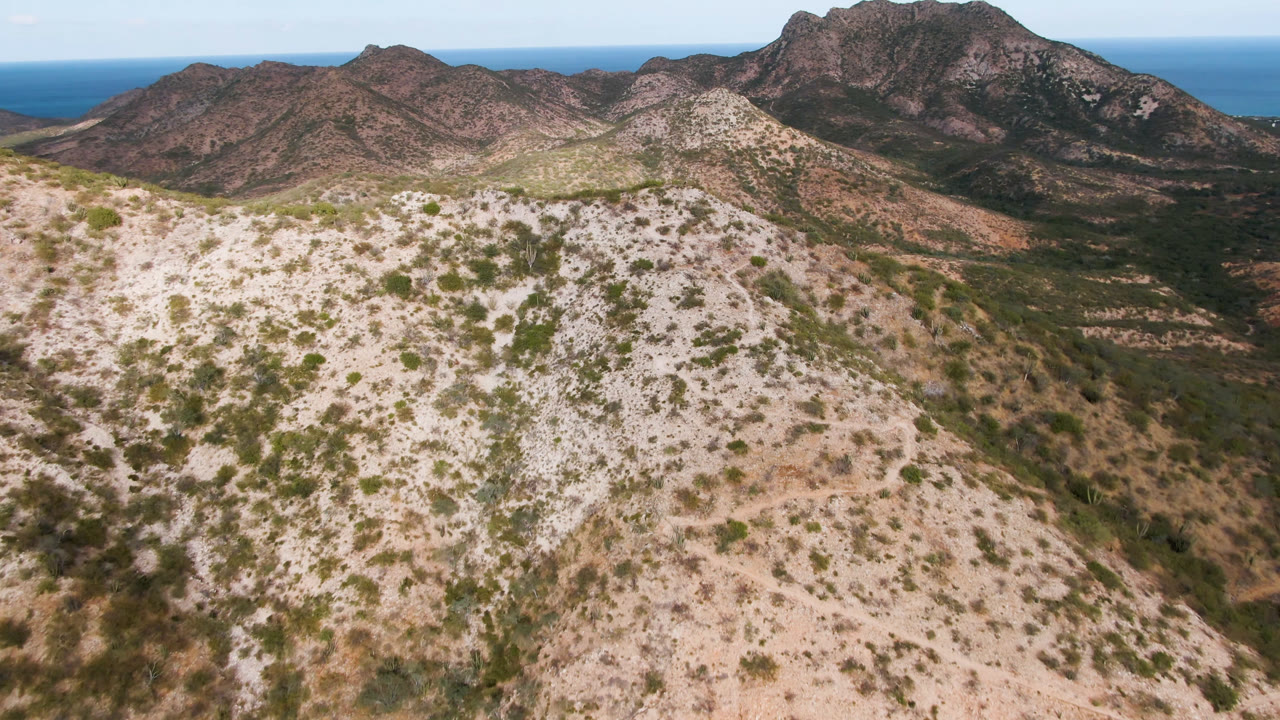 Aerial view in Cabo Pulmo, Baja California Sur, Mexico with a broad pale foreground ridge, scattered desert vegetation, layered brown mountains, distant blue water, and no visible people.