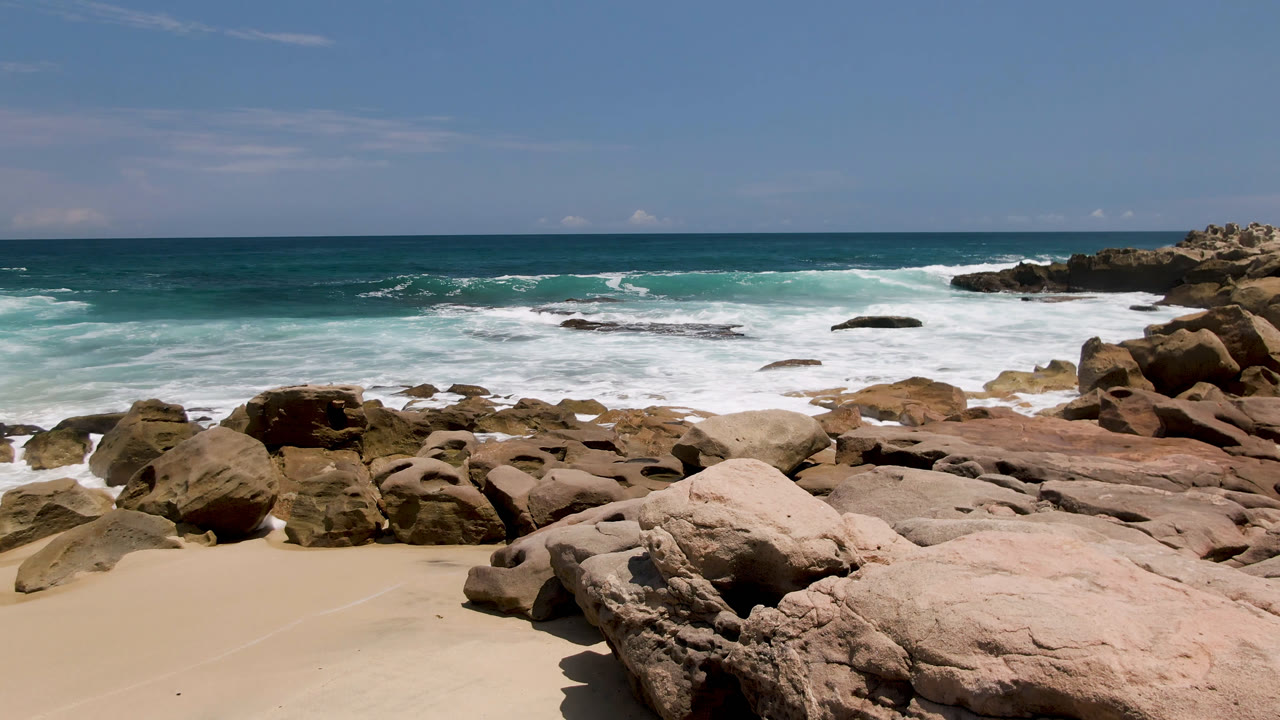 Laguna Beach, Baja California Sur rocky shoreline with tan sand in front, rounded rocks across the foreground, white surf breaking offshore, and no visible people.