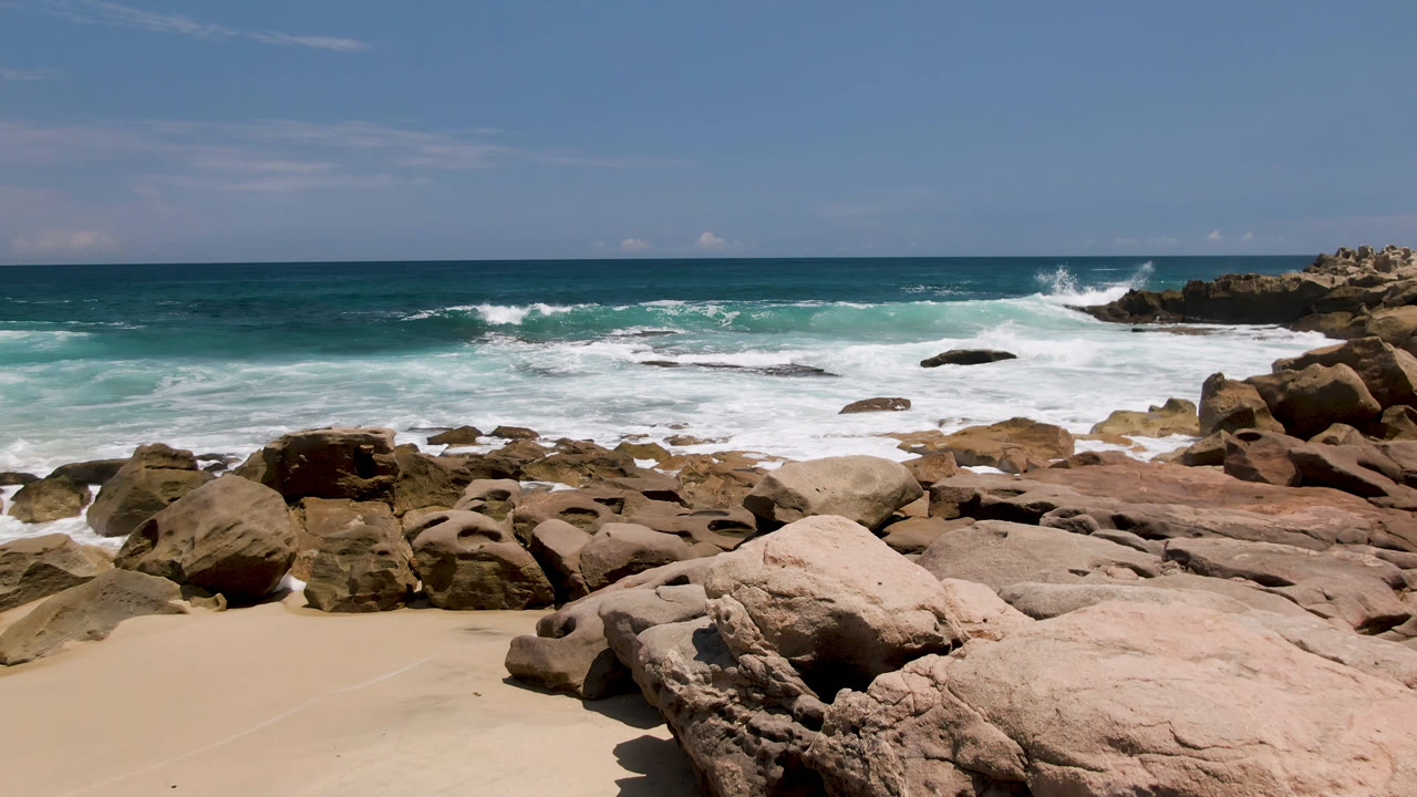 Laguna Beach, Baja California Sur shoreline with foamy surf spreading around offshore rocks, tan boulders in the foreground, and no visible people.