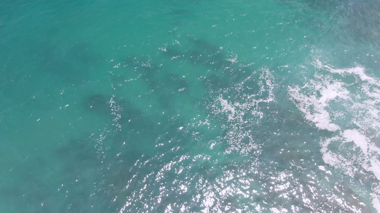 Overhead view of turquoise ocean at Laguna Beach, Baja California Sur, with scattered white surf foam, dark submerged patches, and no visible people.