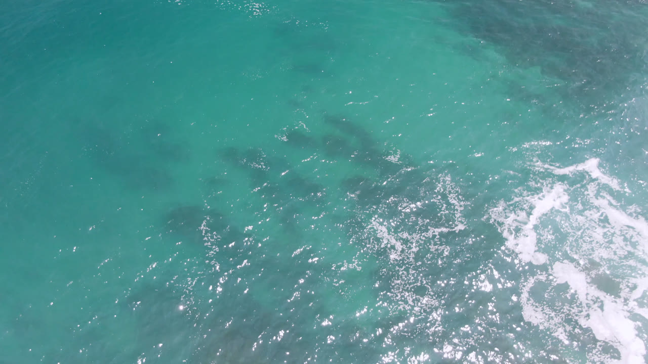 Top-down Laguna Beach ocean view with scattered surf foam, bright reflections on turquoise water, soft darker patches below, and no visible people.