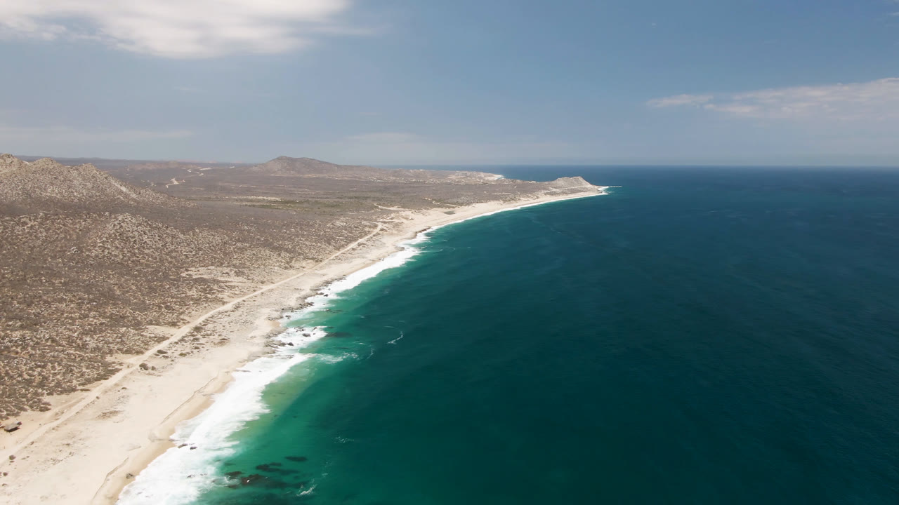 Aerial view of Laguna Beach in Baja California Sur, with pale sand, teal ocean, dry scrubland, distant hills, and no visible people.