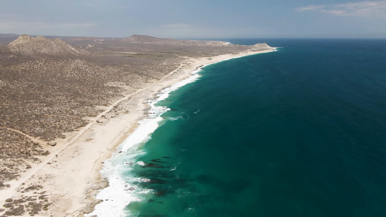 Aerial view over Laguna Beach in Baja California Sur, Mexico, with pale sand, white surf, dry scrubland, rocky hills, and no visible people.
