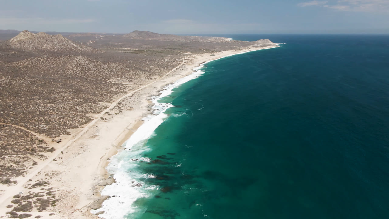 High aerial frame of Laguna Beach, Baja California Sur, showing a long pale shoreline, turquoise shallows, darker offshore water, inland brush, hills, and no visible people.